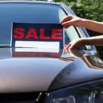 Brownish vehicle with a sign being put on the windshield that says "SALE" in red letters