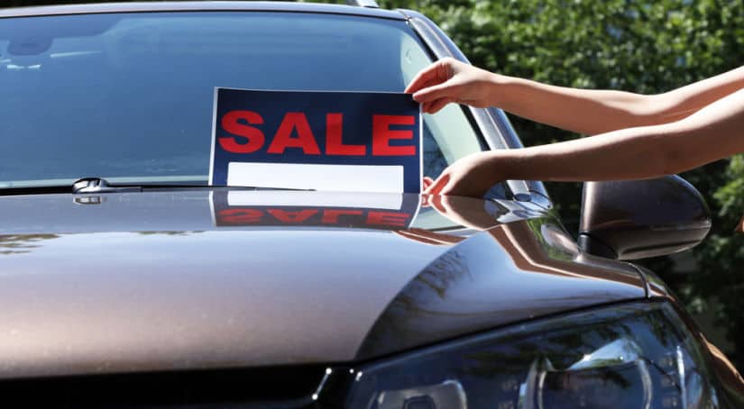 Brownish vehicle with a sign being put on the windshield that says "SALE" in red letters
