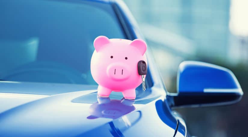Pink piggy bank sitting on the hood of a blue car with a car key attached to it