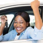 Woman smiling with her arms up, holding a car key, while sitting in a white vehicle's driver's seat
