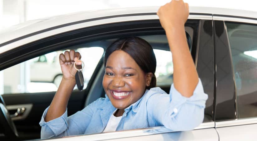 Woman smiling with her arms up, holding a car key, while sitting in a white vehicle's driver's seat