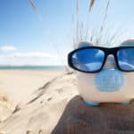 White and blue piggy bank in black and blue sunglasses sitting on a mound of sand with the ocean in the background