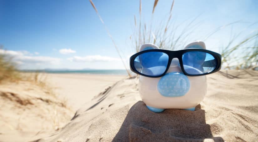 White and blue piggy bank in black and blue sunglasses sitting on a mound of sand with the ocean in the background