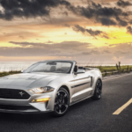 A silver 2019 Ford Mustang convertible with the top down cruises down a sunset lit road
