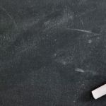 White piece of chalk sitting on a black chalkboard with white dust on it