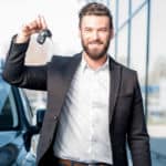 Man in a white shirt and black jacket standing in front of a black car and holding up car keys