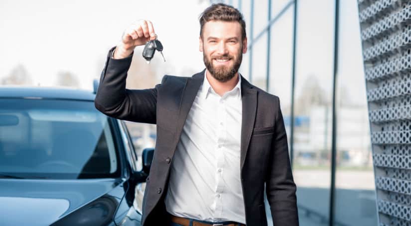 Man in a white shirt and black jacket standing in front of a black car and holding up car keys
