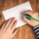 White paper sitting on a brown table being held down by one hand and written on by green pencil held in the other hand