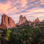 Green trees in front of brown rock mountains with blue and pink cloudy sky in the background