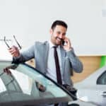 Businessman talking on phone near car in modern dealership.
