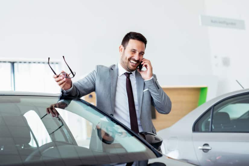 Businessman talking on phone near car in modern dealership.