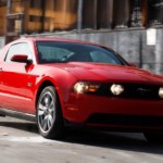 A red 2015 Ford Mustang drives past an industrial building