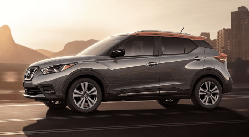 A grey 2019 Nissan Kicks with copper accents driving at sunrise with hills and buildings in the background. Check out the Kicks at a Nissan Dealer.