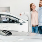 A couple is looking at used cars at a dealership. The woman is pointing to a light silver car.