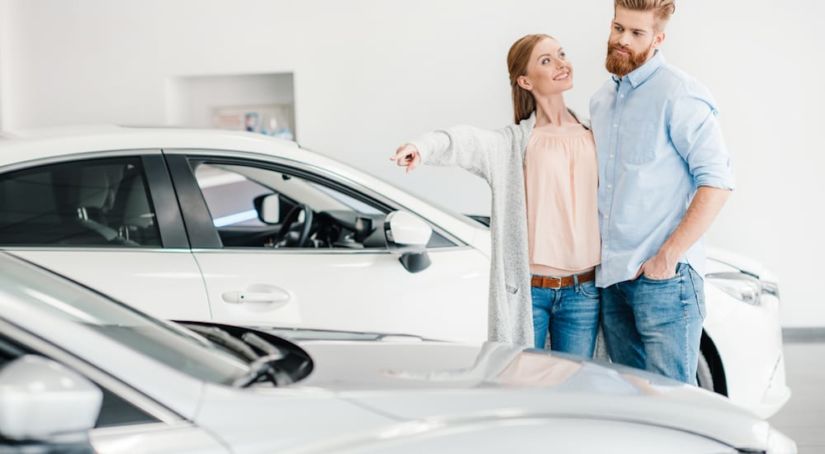 A couple is looking at used cars at a dealership. The woman is pointing to a light silver car.