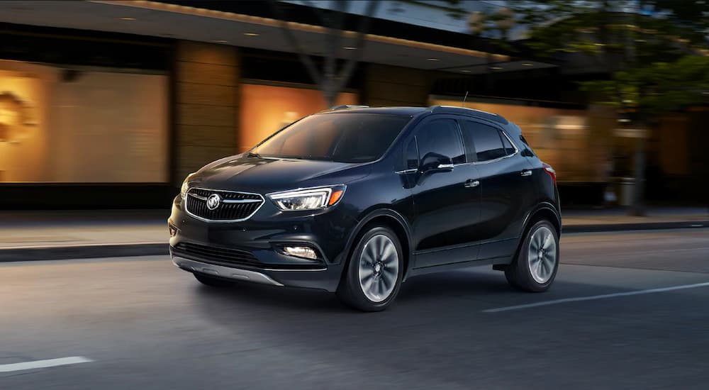 A black 2019 Buick Encore is driving past a building at dusk. Check out handling when comparing the 2019 Buick Encore vs 2019 Nissan Kicks.