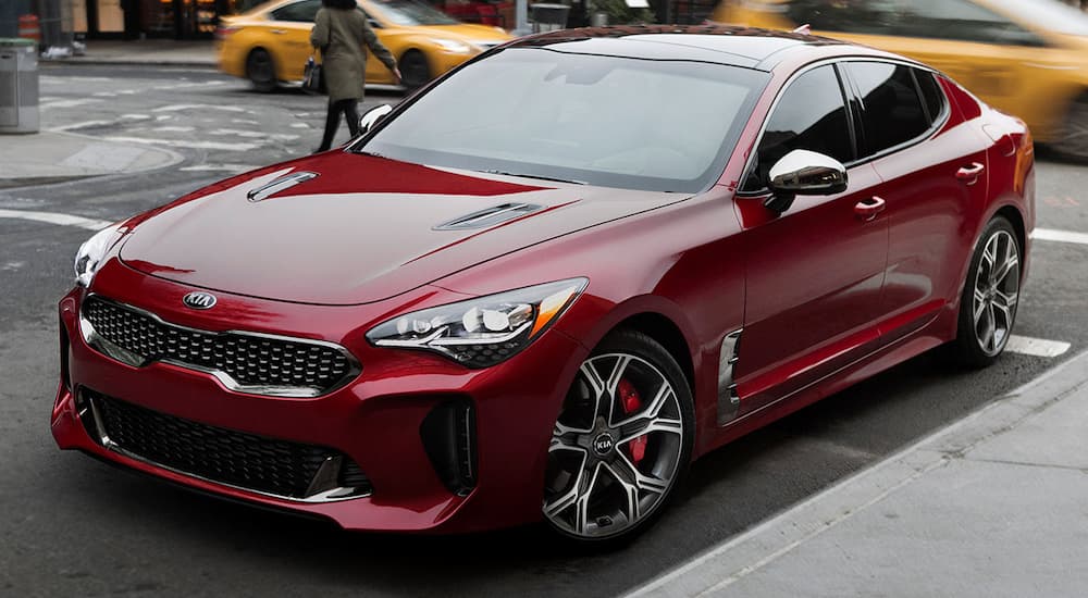 A dark red 2019 Kia Stinger is parked in a city with taxis behind it. If you want to check one out search for 'KIA dealership near me'.