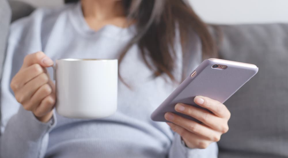 A woman in a sweater is holding a coffee mug and her phone. She is looking up no credit car loans.