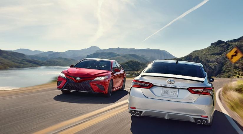 A red and a silver 2019 Toyota Camry are passing each other on a road near a lake and mountains. The Camry is one of the top vehicles purchased at dealerships in Los Angeles.