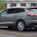 A grey 2019 Buick Enclave is driving in front of buildings.