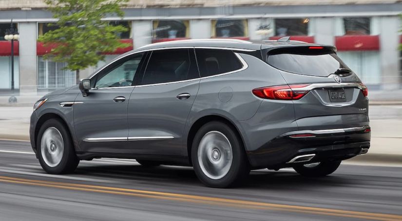 A grey 2019 Buick Enclave is driving in front of buildings.