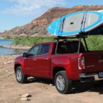 A red 2019 GMC Canyon is parked at a lake with paddle boards on the bed rack.