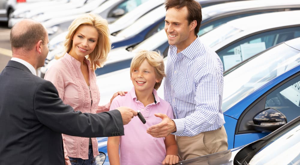 A younger teen boy is with his parents and a salesman looking at cars at a Buick dealership. 