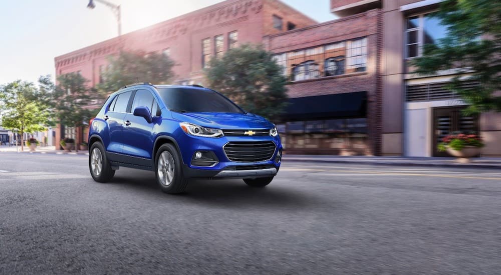 A blue 2017 Chevy Trax, purchased as a Chevy Certified Pre Owned vehicle, is driving in front of brick buildings.