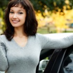 A young woman is leaning on her white car smiling while holding her keys.