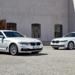Two white 2017 BMW 5 Series are parked in front of a tan building.