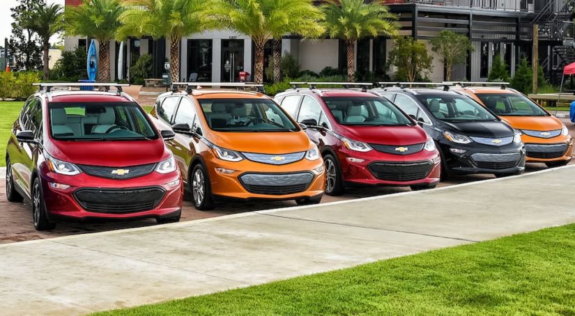 Two red, two orange, and a black 2017 Chevy Bolt EV are in line along a sidewalk.
