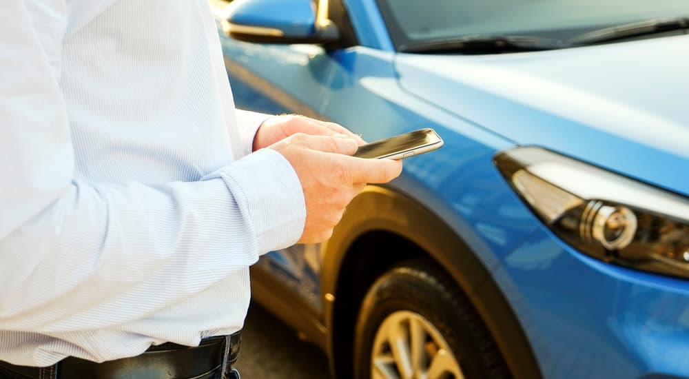 A man is using his smartphone while standing next to a blue car looking up current auto news.