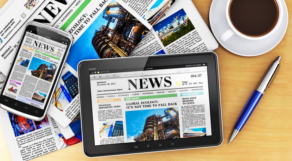 A phone, laptop, and newspaper displaying news headlines are on a table next to a coffee cup.