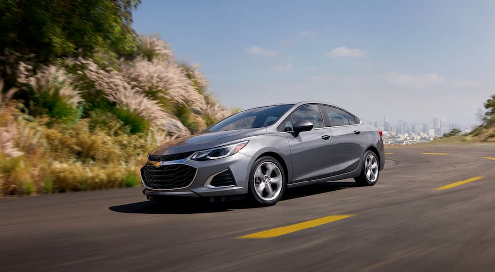 A grey 2019 Chevy Cruze is driving around a corner with a city skyline in the distance.