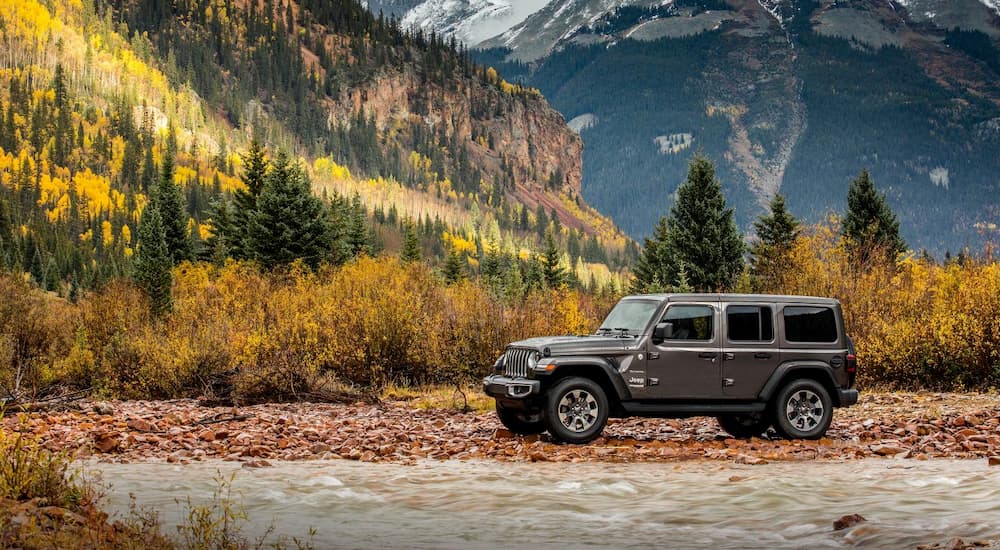A grey 2018 Jeep Wrangler, popular among used SUVs for sale, is on a river bank in front of woods and mountains near Colorado Springs.