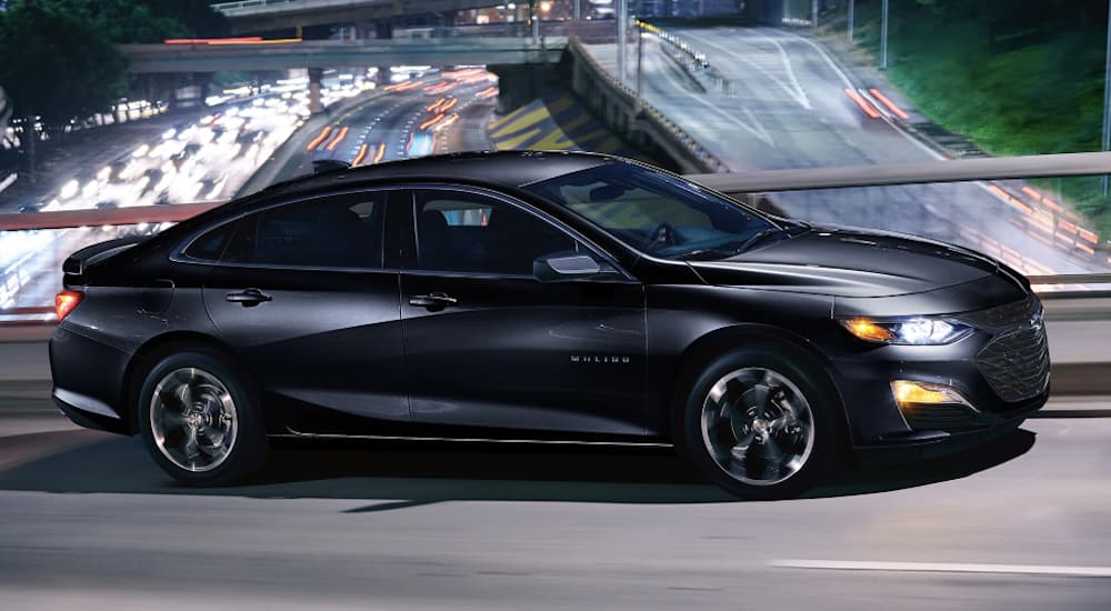 A black 2019 Chevy Malibu is driving on an overpass over a highway at night.