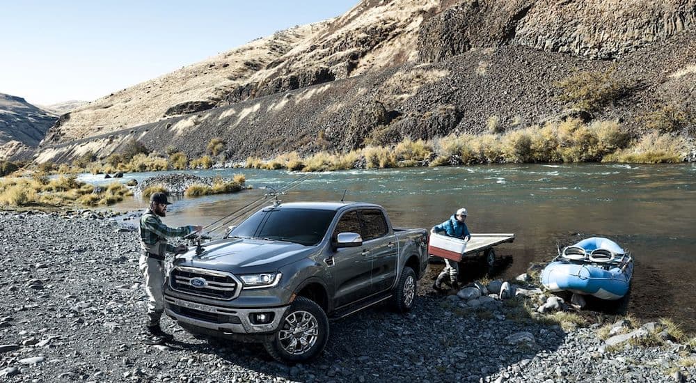 A grey 2019 Ford Ranger is parked near a lake with mountains in the background, with two men taking kayaks out of the bed, 