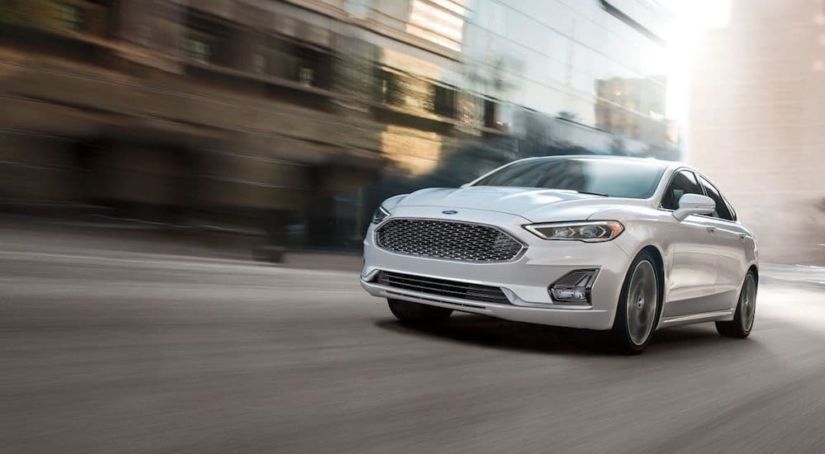 A white 2019 Ford Fusion, popular among Ford models, driving down a city street with blurred buildings in the background.
