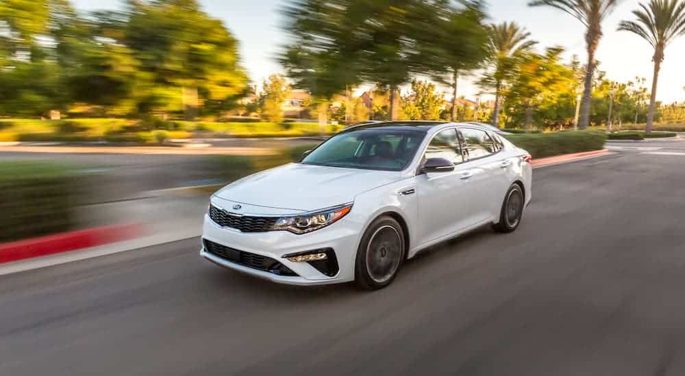 A white 2020 Kia Optima is driving down a city street during the daytime. 