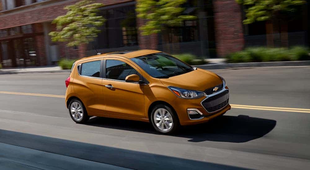 An orange 2020 Chevy Spark is driving past brick buildings.