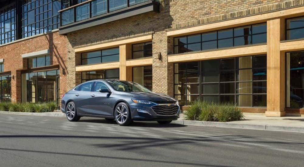 A grey 2019 Chevy Malibu Hybrid, with one of our Top 15 / Automotive Features, is parked in front of a brick building. 