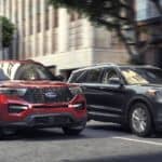 Two 2020 Ford Explorers, red and grey, are stopped at a red light on a busy city street.
