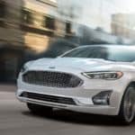A white 2020 Ford Fusion is driving on a city street.