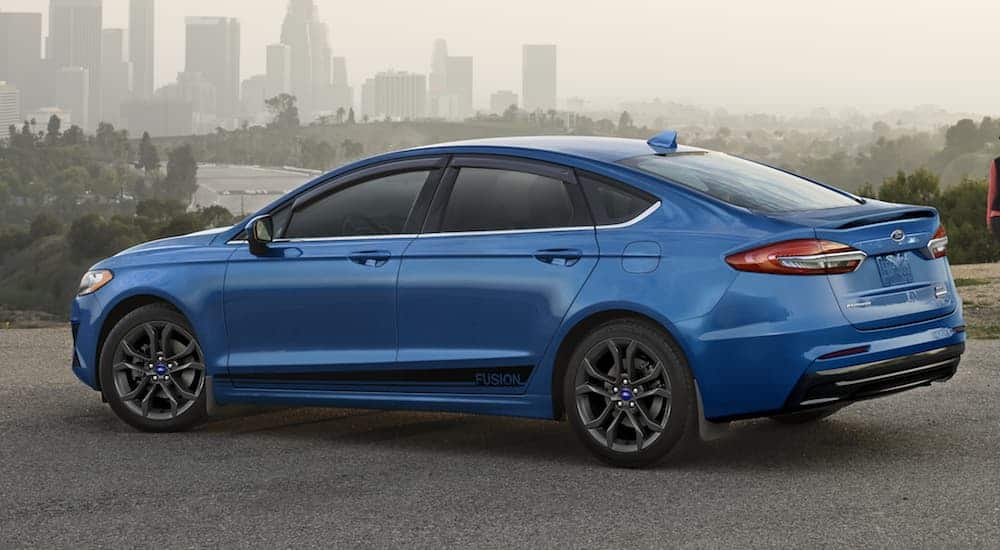 A blue 2020 Ford Fusion is parked on a road with a city skyline in the distance. 