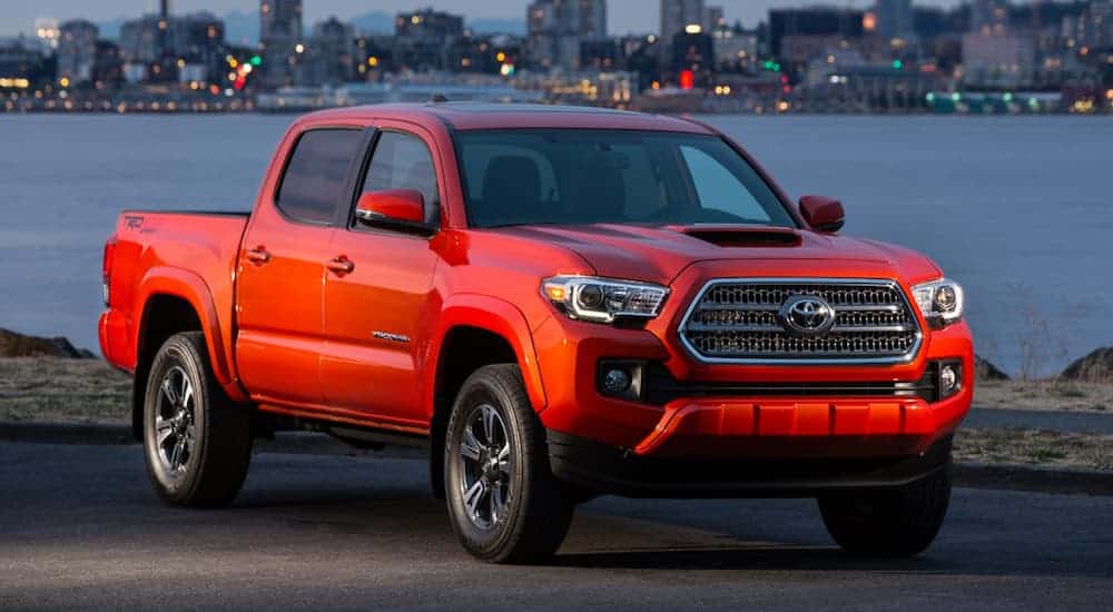 A red 2016 Toyota Tacoma is parked in front of a lake with city lights in the distance. 