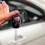 A hand is holding a car key with a silver car in the background.