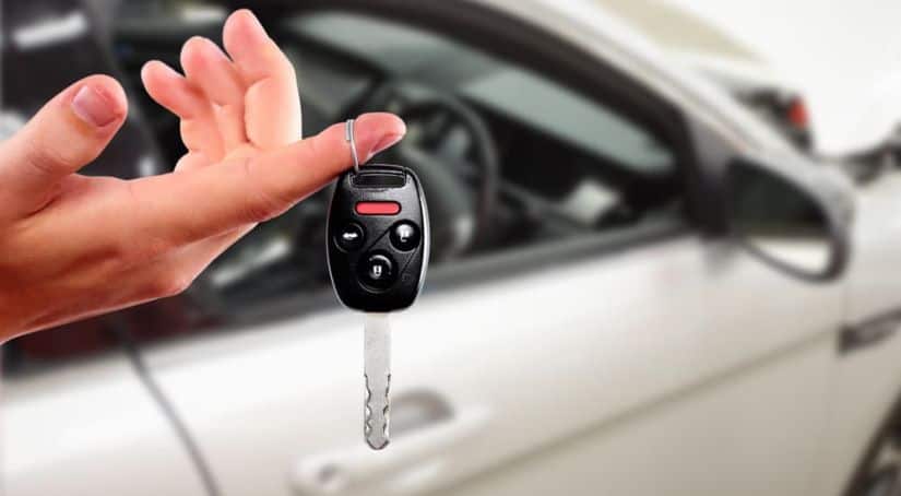 A hand is holding a car key with a silver car in the background.
