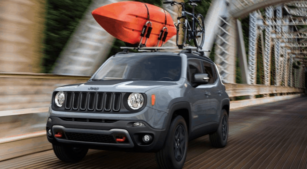 A grey 2020 Jeep Renegade is driving on a bridge with a kayak and a bike on the roof racks. 