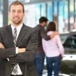 A smiling salesman has his arms crossed while standing in a showroom at a Chevy dealer.