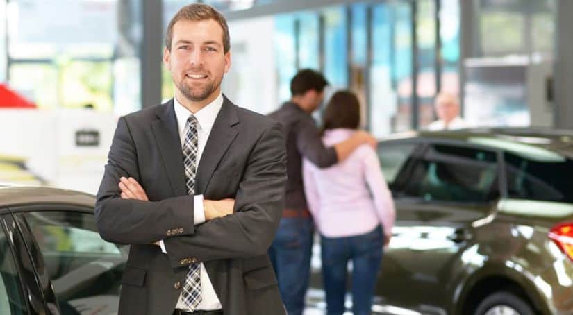 A smiling salesman has his arms crossed while standing in a showroom at a Chevy dealer.